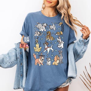 May include: Blue jean t-shirt featuring a collection of cartoon dog illustrations. The dogs are in various poses and styles, with blue hearts scattered around them. The shirt has rolled-up sleeves.