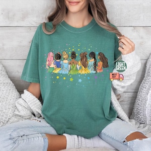 Puede incluir: Una camiseta verde con un gr&aacute;fico de siete princesas de dibujos animados de pie juntas. Las princesas llevan vestidos coloridos y tienen diferentes peinados. El gr&aacute;fico est&aacute; rodeado de estrellas amarillas.
