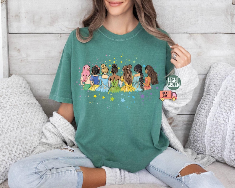 Puede incluir: Una camiseta verde con un gr&aacute;fico de siete princesas de dibujos animados de pie juntas. Las princesas llevan vestidos coloridos y tienen diferentes peinados. El gr&aacute;fico est&aacute; rodeado de estrellas amarillas.