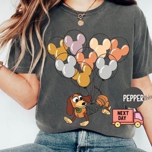 May include: A gray t-shirt with a cartoon image of a dog holding a bunch of Mickey Mouse-shaped balloons. The balloons are in shades of orange, yellow, pink, and gray. The text "NEXT DAY" is printed on a small truck in the bottom right corner of the image.