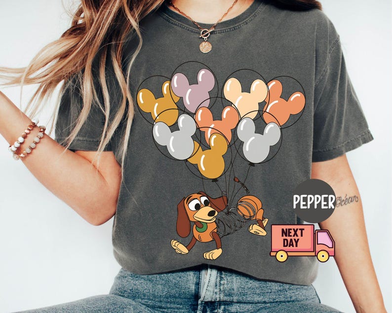 May include: A gray t-shirt with a cartoon image of a dog holding a bunch of Mickey Mouse-shaped balloons. The balloons are in shades of orange, yellow, pink, and gray. The text "NEXT DAY" is printed on a small truck in the bottom right corner of the image.