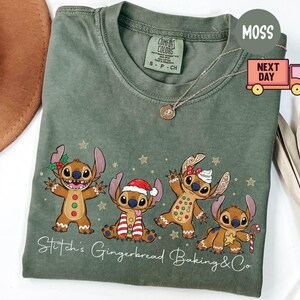 Puede incluir: Camiseta verde salvia con un dise&ntilde;o festivo de cuatro personajes de estilo pan de jengibre que se asemejan a Stitch. Los personajes est&aacute;n decorados con accesorios navide&ntilde;os y estrellas doradas. El texto "Stitch's Gingerbread Baking & Co" est&aacute; impreso en la parte inferior.