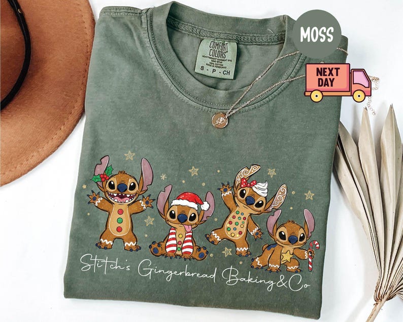 Puede incluir: Camiseta verde salvia con un dise&ntilde;o festivo de cuatro personajes de estilo pan de jengibre que se asemejan a Stitch. Los personajes est&aacute;n decorados con accesorios navide&ntilde;os y estrellas doradas. El texto "Stitch's Gingerbread Baking & Co" est&aacute; impreso en la parte inferior.