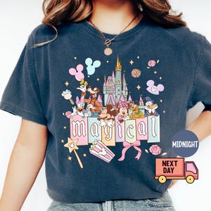 May include: Dark blue t-shirt featuring a colorful graphic with Disney characters, a castle, and the word "magical". The design includes Mickey Mouse, Donald Duck, and other characters, with stars and flowers. A magic wand and popcorn are also included.