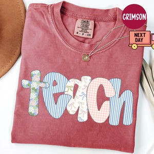 May include: A red Comfort Colors t-shirt with the word "TEACH" in a patchwork design. The letters are in floral, gingham, and striped patterns. A gold necklace is draped over the shirt.
