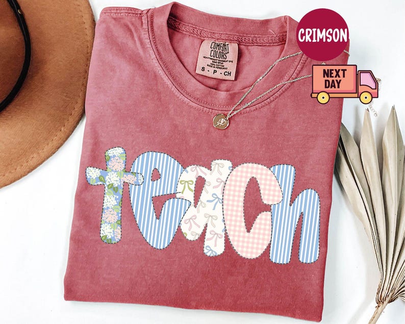 May include: A red Comfort Colors t-shirt with the word "TEACH" in a patchwork design. The letters are in floral, gingham, and striped patterns. A gold necklace is draped over the shirt.