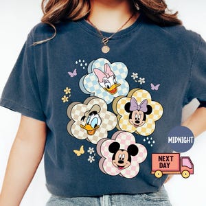 May include: Dark blue t-shirt featuring a floral design with cartoon characters Daisy Duck, Donald Duck, Minnie Mouse, and Mickey Mouse. The flowers have checkered patterns in pastel colors. Butterflies and small flowers are also present.