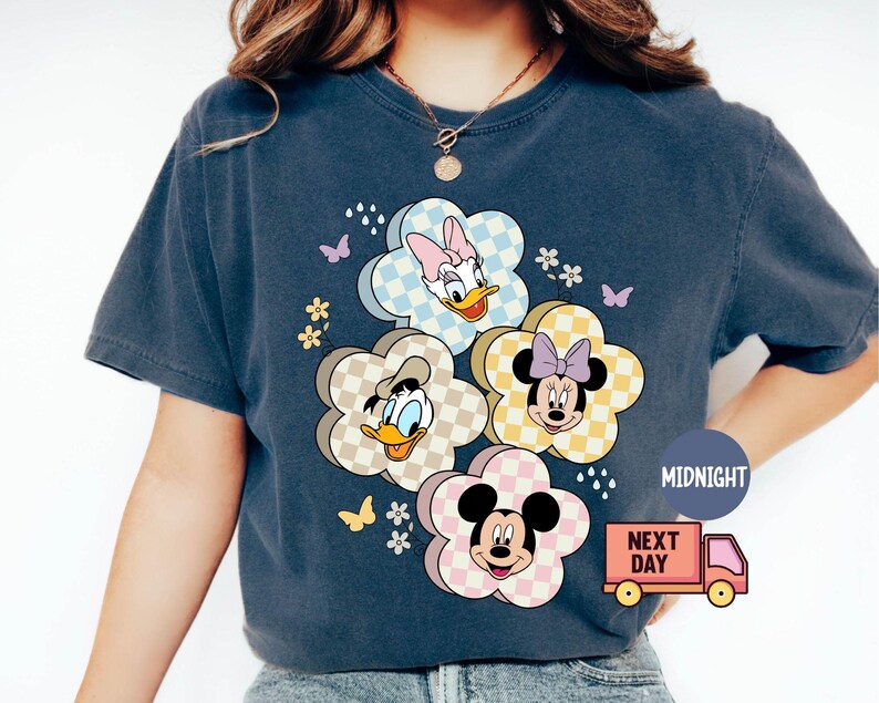 May include: Dark blue t-shirt featuring a floral design with cartoon characters Daisy Duck, Donald Duck, Minnie Mouse, and Mickey Mouse. The flowers have checkered patterns in pastel colors. Butterflies and small flowers are also present.