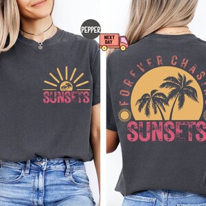 May include: Dark gray t-shirt with a sunset graphic on the front and back. The front features a sun and palm trees with the word "SUNSETS". The back says "FOREVER CHASING SUNSETS" with a similar design.