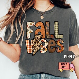 May include: A grey t-shirt with a patchwork design that says "Fall Vibes". The design features a lightning bolt with a leopard print, plaid, and autumn leaves.