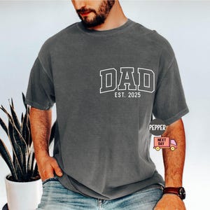 May include: Dark gray t-shirt with the word "DAD" in white block letters and "EST. 2025" underneath. The shirt is a relaxed fit and appears to be made of a soft material. The shirt is worn with blue jeans.