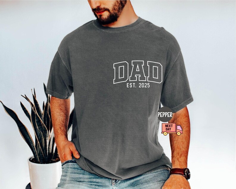 May include: Dark gray t-shirt with the word "DAD" in white block letters and "EST. 2025" underneath. The shirt is a relaxed fit and appears to be made of a soft material. The shirt is worn with blue jeans.