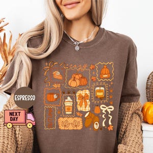 May include: Brown t-shirt with a fall-themed graphic. The design includes pumpkins, a slice of pie, a coffee cup, a hat, and the word "cozy" in a decorative frame. The shirt is a warm, earthy tone, perfect for autumn.