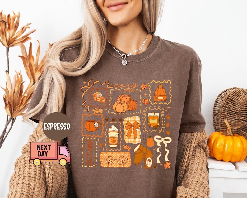 May include: Brown t-shirt with a fall-themed graphic. The design includes pumpkins, a slice of pie, a coffee cup, a hat, and the word "cozy" in a decorative frame. The shirt is a warm, earthy tone, perfect for autumn.