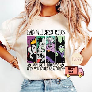May include: Ivory-colored t-shirt featuring the text "BAD WITCHES CLUB" above four cartoon villain portraits. Below, the shirt reads "WHY BE A PRINCESS WHEN YOU COULD BE A QUEEN?" A small pumpkin graphic is on either side of the text.