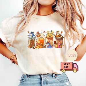 May include: Ivory-colored t-shirt featuring a colorful graphic of Winnie the Pooh characters in fall-themed coffee drinks. The design includes Eeyore, Piglet, Pooh, and Tigger, with pumpkins and autumn leaves. The shirt has a crew neck and short sleeves.