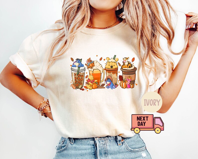 May include: Ivory-colored t-shirt featuring a colorful graphic of Winnie the Pooh characters in fall-themed coffee drinks. The design includes Eeyore, Piglet, Pooh, and Tigger, with pumpkins and autumn leaves. The shirt has a crew neck and short sleeves.