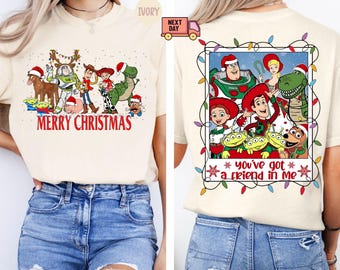 May include: A white t-shirt with a Christmas-themed image of the Toy Story characters. The image features Woody, Buzz Lightyear, Jessie, and other characters from the movie. The text "Merry Christmas" is printed on the shirt. The second image shows a white t-shirt with a picture of the Toy Story characters in a frame with Christmas lights. The text "You've got a friend in me" is printed on the shirt.