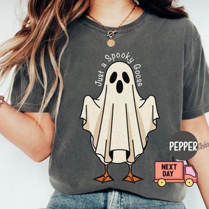 May include: Dark gray t-shirt featuring a cartoon ghost with goose feet. The ghost has a white body, black eyes, and a beak. The text "Just a Spooky Goose" is arched above the ghost. The shirt is a comfortable fit.
