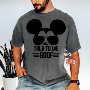 May include: A charcoal gray short-sleeve t-shirt featuring a black graphic of a mouse silhouette wearing sunglasses. The text "TALK TO ME GOOF" is printed in black block letters below the sunglasses, flanked by two horizontal lines on each side.