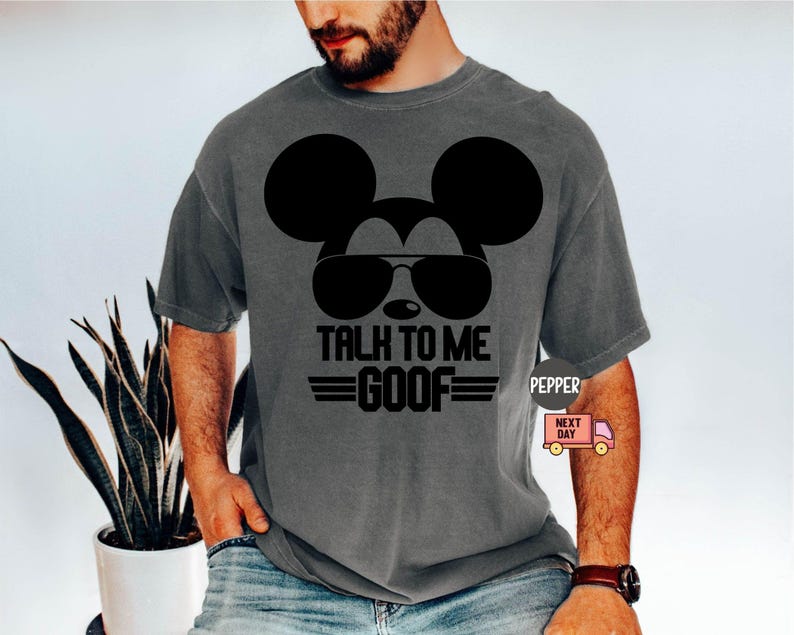 May include: A charcoal gray short-sleeve t-shirt featuring a black graphic of a mouse silhouette wearing sunglasses. The text "TALK TO ME GOOF" is printed in black block letters below the sunglasses, flanked by two horizontal lines on each side.