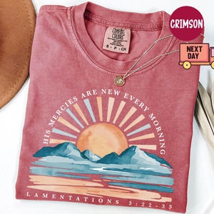 Könnte beinhalten: Ein staubrosa Comfort Colors T-Shirt mit einer Grafik eines Sonnenaufgangs über Bergen und dem Text "HIS MERCIES ARE NEW EVERY MORNING" und "LAMENTATIONS 3:22-33". Eine goldene Halskette mit Anhänger befindet sich auf dem Shirt.