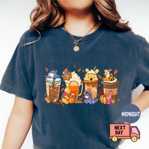 May include: Dark blue t-shirt featuring a colorful graphic of Winnie the Pooh characters as coffee drinks with fall-themed decorations. The design includes pumpkins and autumn leaves. The shirt has a relaxed fit.