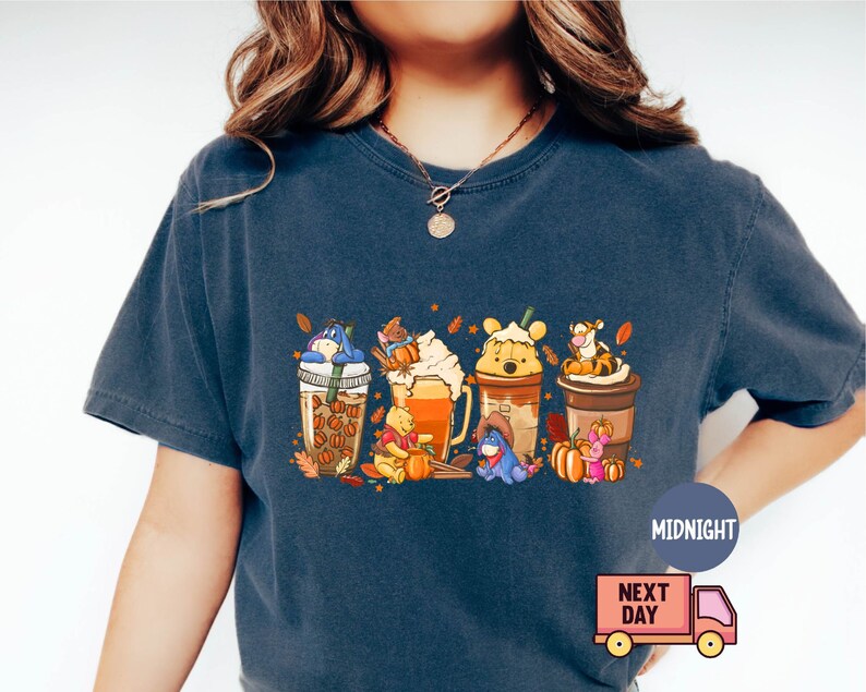 May include: Dark blue t-shirt featuring a colorful graphic of Winnie the Pooh characters as coffee drinks with fall-themed decorations. The design includes pumpkins and autumn leaves. The shirt has a relaxed fit.