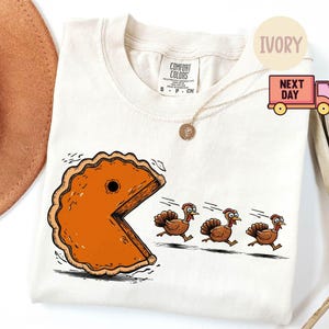 Puede incluir: Camiseta de color marfil con un gráfico humorístico de una porción de pastel que se asemeja a Pac-Man persiguiendo a tres pavos. El diseño es en tonos naranja, marrón y negro. La camiseta tiene cuello redondo y mangas cortas.