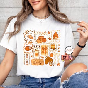 May include: White t-shirt featuring a collage of autumn-themed illustrations in orange and brown. The design includes pumpkins, a slice of pie, a mug, a latte, a candle, a bow, a hat, and leaves. The words "cozy" and "autumn" are also included.