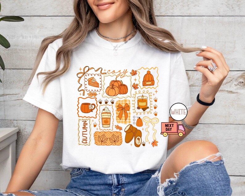 May include: White t-shirt featuring a collage of autumn-themed illustrations in orange and brown. The design includes pumpkins, a slice of pie, a mug, a latte, a candle, a bow, a hat, and leaves. The words "cozy" and "autumn" are also included.