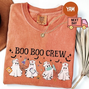 May include: Orange Comfort Colors t-shirt with the text "BOO BOO CREW" and a graphic of ghost characters dressed as medical professionals. The design includes bats, stars, and a pumpkin, perfect for Halloween.