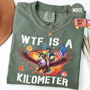 May include: Olive green t-shirt with the text "WTF IS A KILOMETER" in white. The graphic depicts a soldier riding an eagle, holding a rifle and an American flag, with a colorful explosion background.