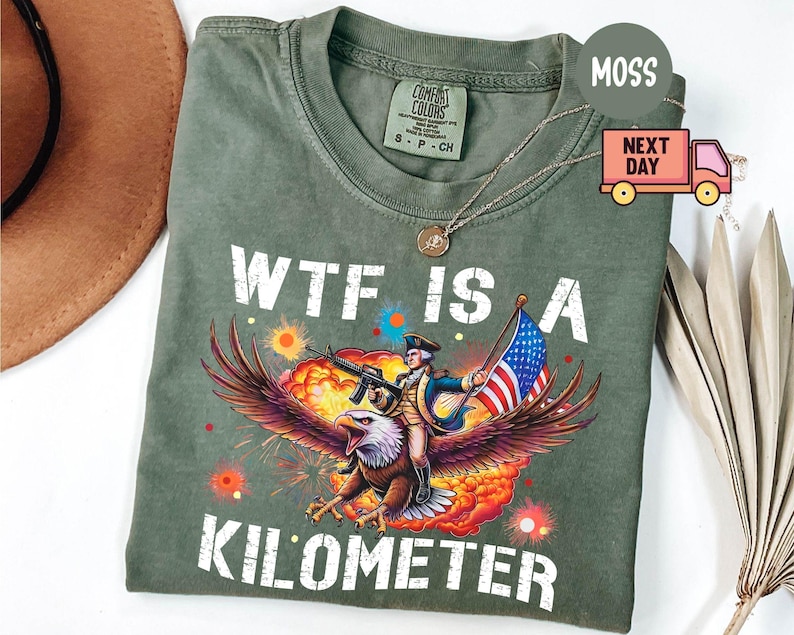 May include: Olive green t-shirt with the text "WTF IS A KILOMETER" in white. The graphic depicts a soldier riding an eagle, holding a rifle and an American flag, with a colorful explosion background.