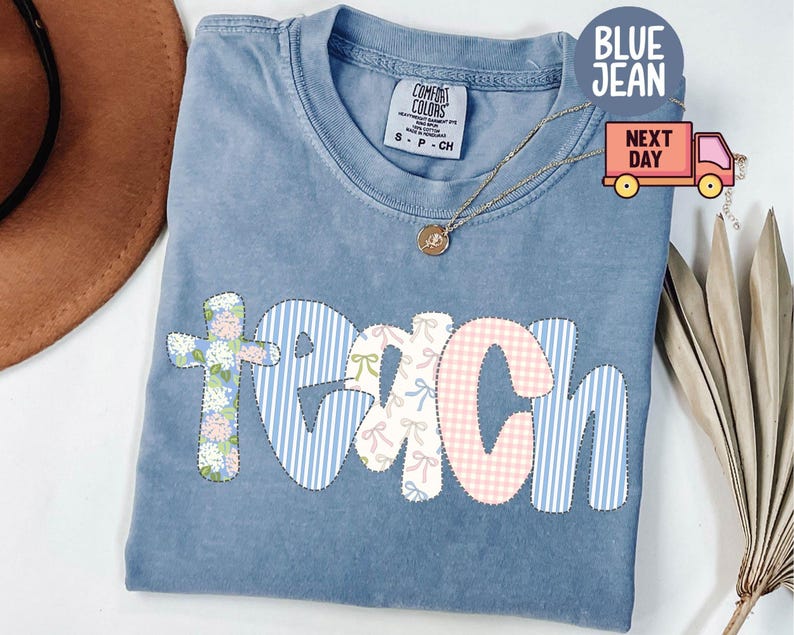 May include: A blue Comfort Colors t-shirt with the word "TEACH" appliqued on the front. The letters are in various patterns, including floral, blue stripes, and pink gingham. A gold necklace with a pendant is draped on the shirt.