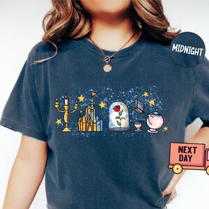 May include: A dark blue t-shirt with a white and yellow graphic print of a castle, a rose in a glass dome, a candlestick, a teacup, and a teapot. The graphic is on a blue background with white speckles. The shirt has a crew neck and short sleeves.