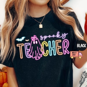 May include: Black t-shirt with a neon graphic that reads "spooky TEACHER" with a ghost and bat design. The shirt has a round neck and short sleeves. A black circle with the word "BLACK" is on the right side of the shirt.