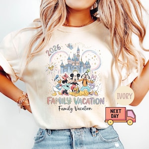 Może przedstawiać: Kremowa koszulka z kolorową grafiką postaci Disneya przed zamkiem, z napisem "2026" i "Family Vacation". Projekt zawiera Myszkę Miki, Minnie, Kaczora Donalda, Daisy Duck i Goofy'ego.
