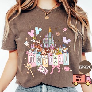 May include: Espresso-colored t-shirt featuring a colorful graphic of Disney characters, a castle, and the word "magical." The design includes Mickey and Minnie Mouse, Donald Duck, and Goofy. A pink bow and popcorn box are also featured.