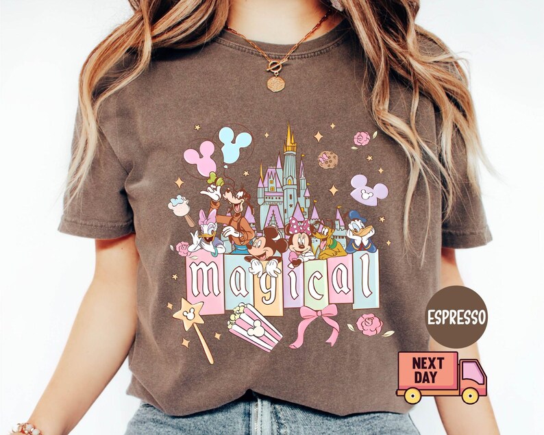 May include: Espresso-colored t-shirt featuring a colorful graphic of Disney characters, a castle, and the word "magical." The design includes Mickey and Minnie Mouse, Donald Duck, and Goofy. A pink bow and popcorn box are also featured.