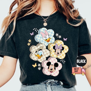 May include: Black t-shirt featuring a floral design with cartoon characters. The flowers have checkered patterns and feature Daisy Duck, Donald Duck, Minnie Mouse, and Mickey Mouse. Butterflies and small flowers are also included in the design.