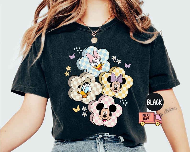 May include: Black t-shirt featuring a floral design with cartoon characters. The flowers have checkered patterns and feature Daisy Duck, Donald Duck, Minnie Mouse, and Mickey Mouse. Butterflies and small flowers are also included in the design.