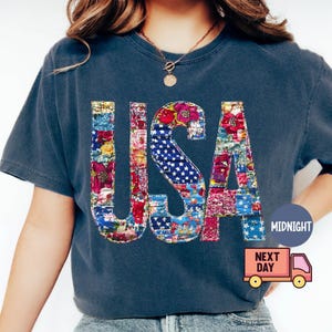 May include: A navy blue t-shirt with the word "USA" in a patchwork design. The letters are filled with floral patterns and a section of the American flag. The shirt has short sleeves and a round neckline.