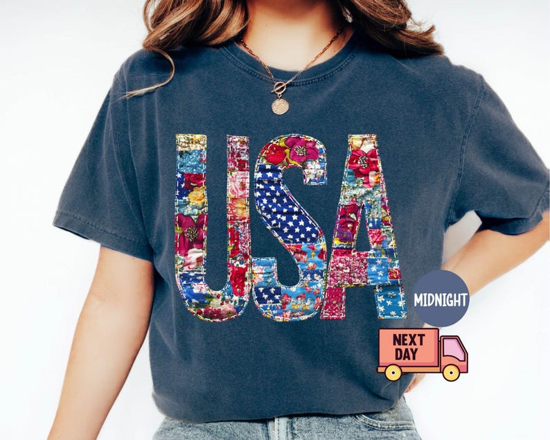 May include: A navy blue t-shirt with the word "USA" in a patchwork design. The letters are filled with floral patterns and a section of the American flag. The shirt has short sleeves and a round neckline.