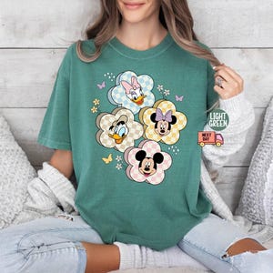 May include: Light green t-shirt featuring a floral design with Disney characters Mickey, Minnie, Donald, and Daisy Duck. The flowers have a checkered pattern. The shirt also has the text "LIGHT GREEN" and a small truck graphic.