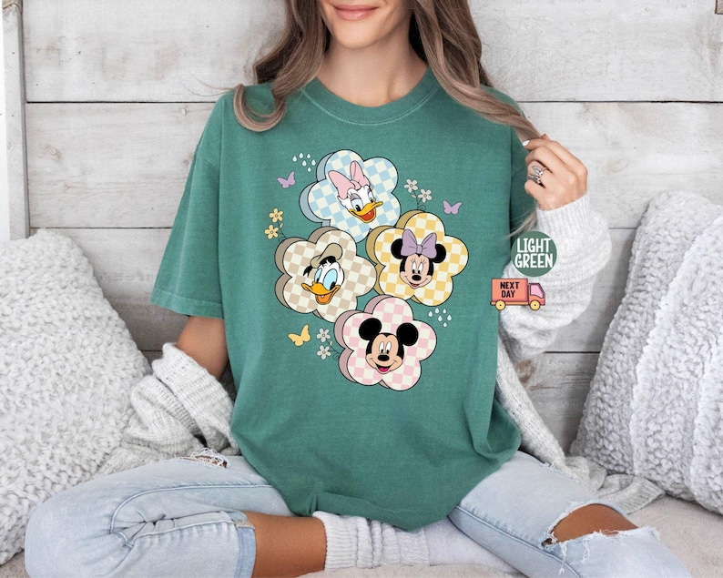 May include: Light green t-shirt featuring a floral design with Disney characters Mickey, Minnie, Donald, and Daisy Duck. The flowers have a checkered pattern. The shirt also has the text "LIGHT GREEN" and a small truck graphic.