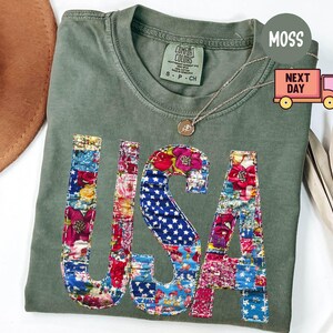 May include: An olive green t-shirt with the word "USA" appliqued on the front in a patchwork design of floral and star-spangled fabrics. The shirt has a crew neck and short sleeves. A gold necklace with a pendant is draped on the shirt.