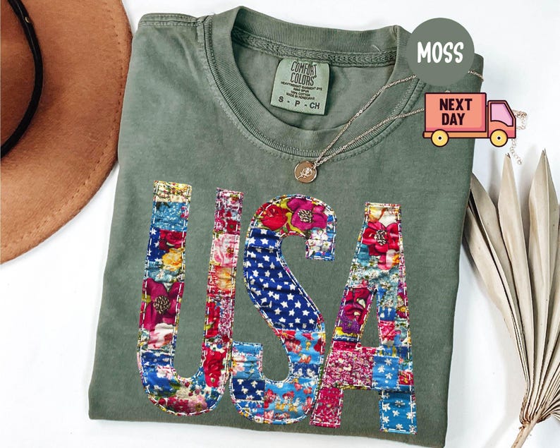 May include: An olive green t-shirt with the word "USA" appliqued on the front in a patchwork design of floral and star-spangled fabrics. The shirt has a crew neck and short sleeves. A gold necklace with a pendant is draped on the shirt.