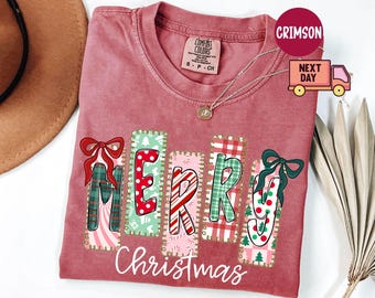 May include: A maroon Comfort Colors t-shirt with the word "MERRY Christmas" in a colorful, festive design. The letters are decorated with bows, candy canes, and plaid patterns, perfect for the holiday season.