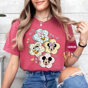 May include: A red t-shirt featuring a floral design with cartoon characters. The flowers have checkered patterns and feature Mickey Mouse, Minnie Mouse, Donald Duck, and Daisy Duck. Small butterflies and flowers are also included in the design.